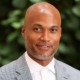 Jermaine Myrie named CEO of MENTOR: black, bald man with greying short beard wearing suit in front of out-of-focus plant backdrop
