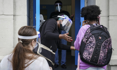 Foster Care Child Removal: Asult in plastic dac shield and face mask stands in doorway gesturing to dark-haired person with multi-colored backpack while another person waits in line