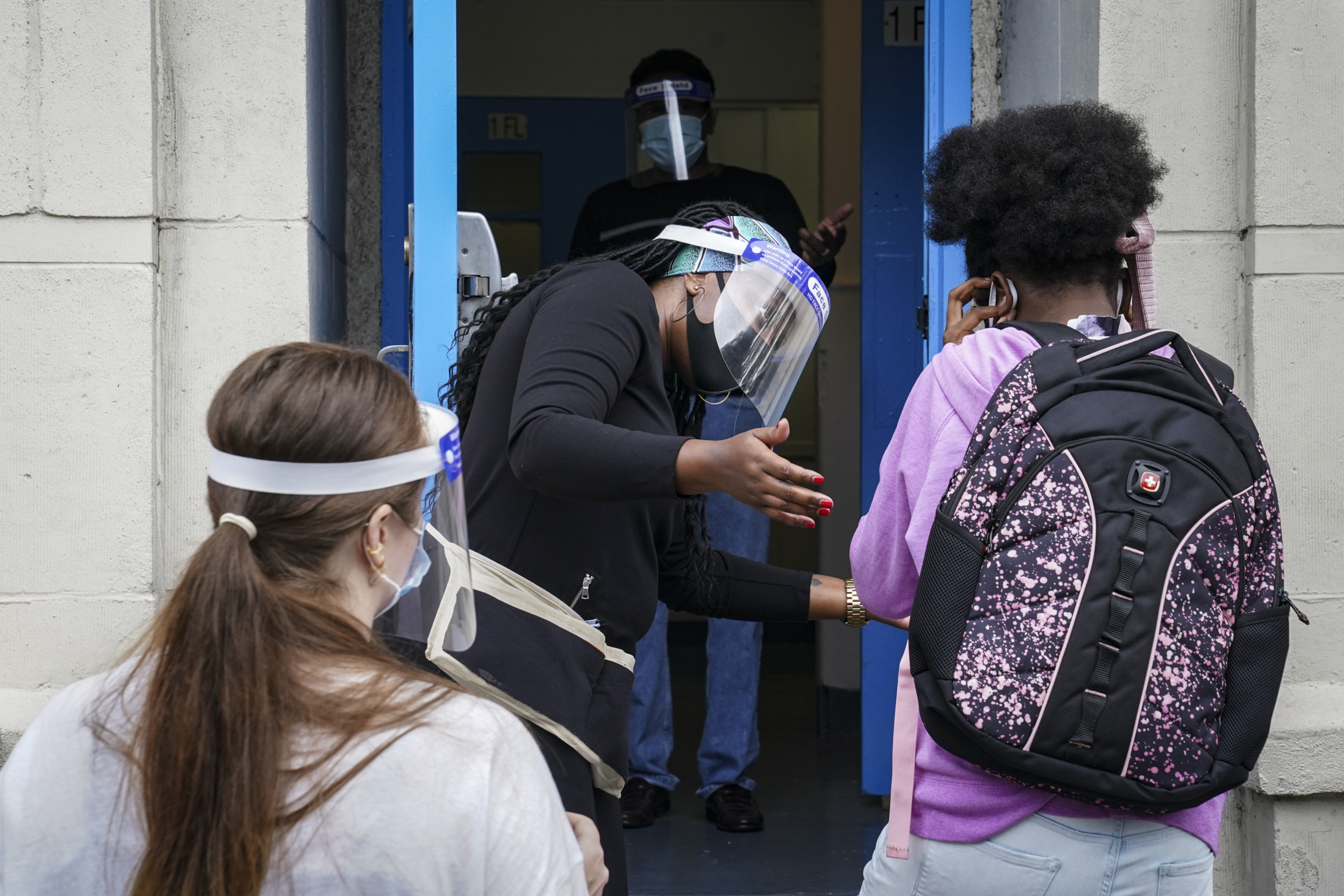 Foster Care Child Removal: Asult in plastic dac shield and face mask stands in doorway gesturing to dark-haired person with multi-colored backpack while another person waits in line