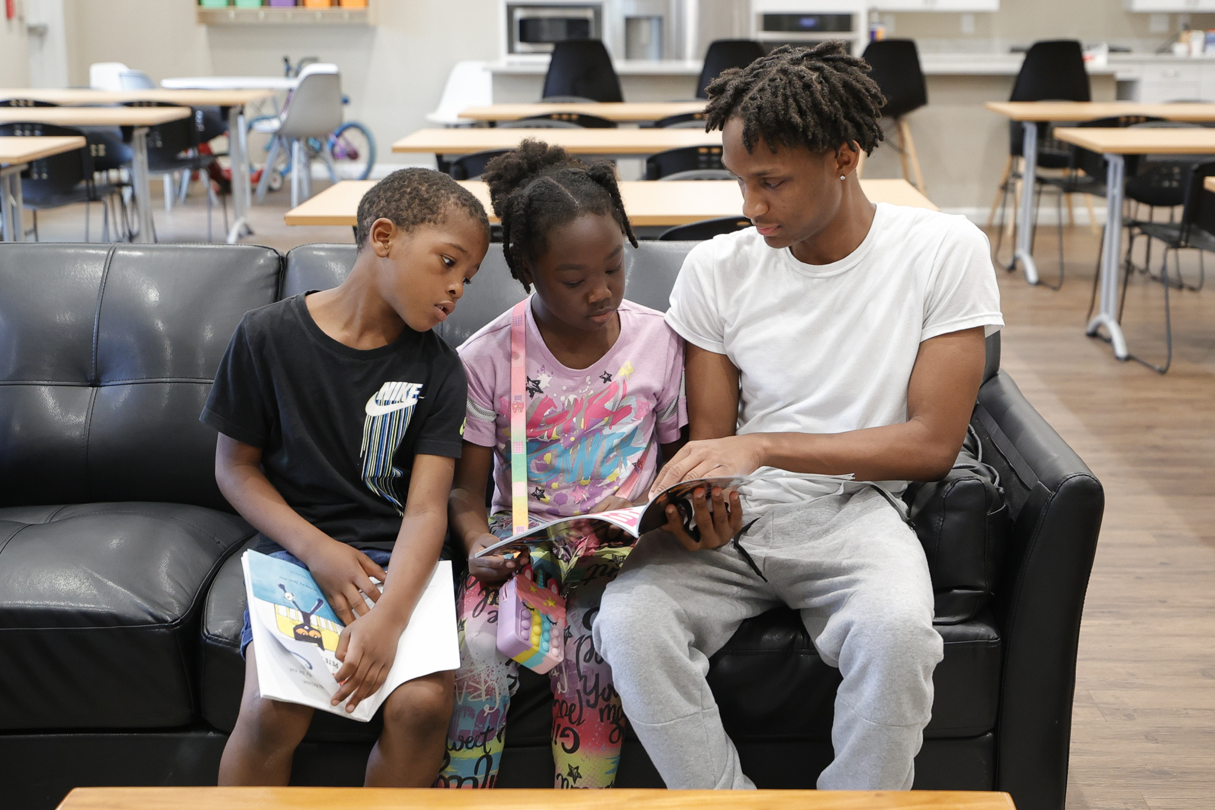 Reading: Two young Black children sit on couch holding books sitting next to Black adult helping them read