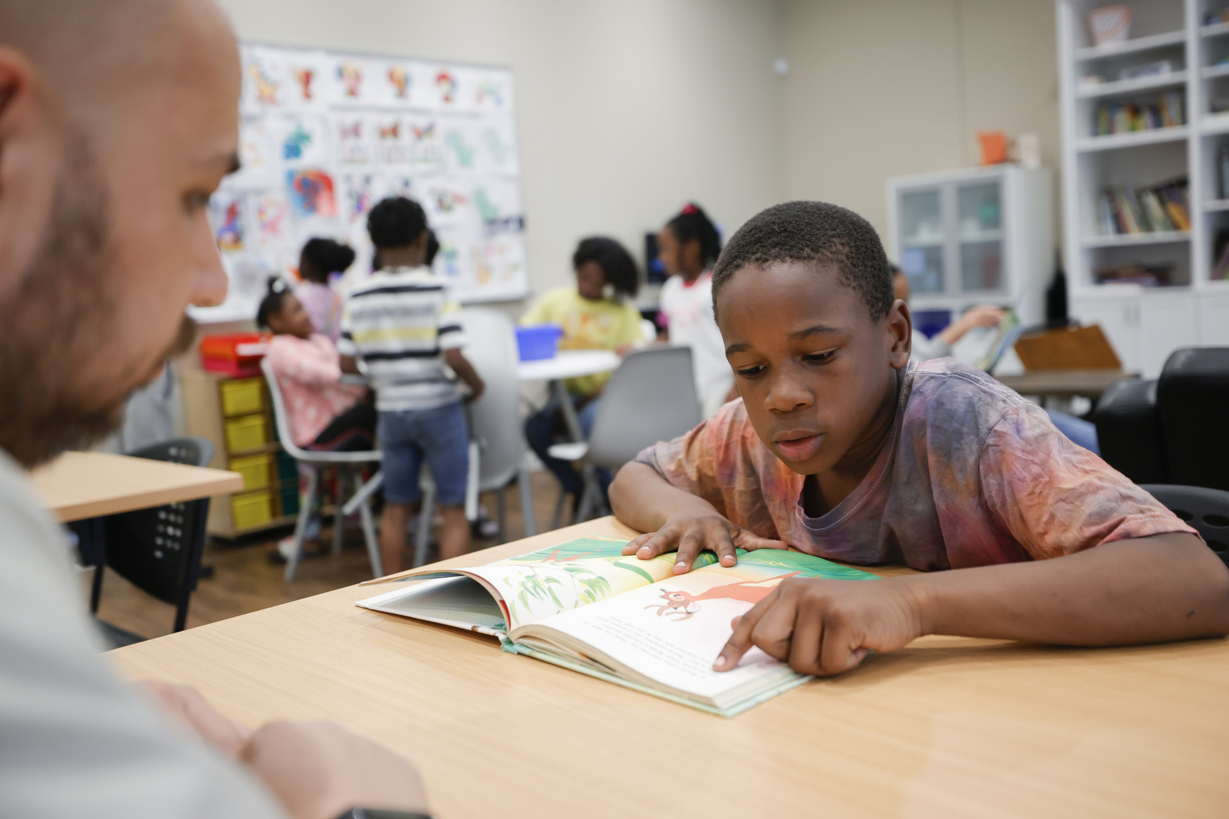 Reading: Young black boy sits at desk leaning forward with finger pointing to word in open workbook.