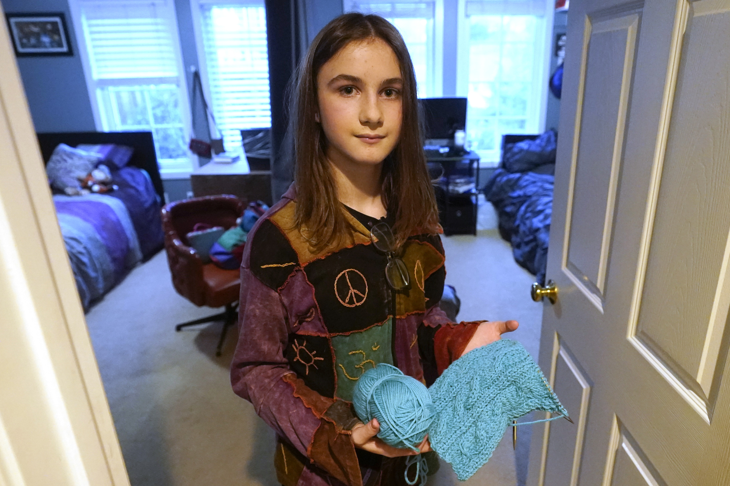 Transgender treatment for kids: Young person with long. dark hair in dark top holdsball and skein of bright turquoise yarn while standing in doorway to a bedroom