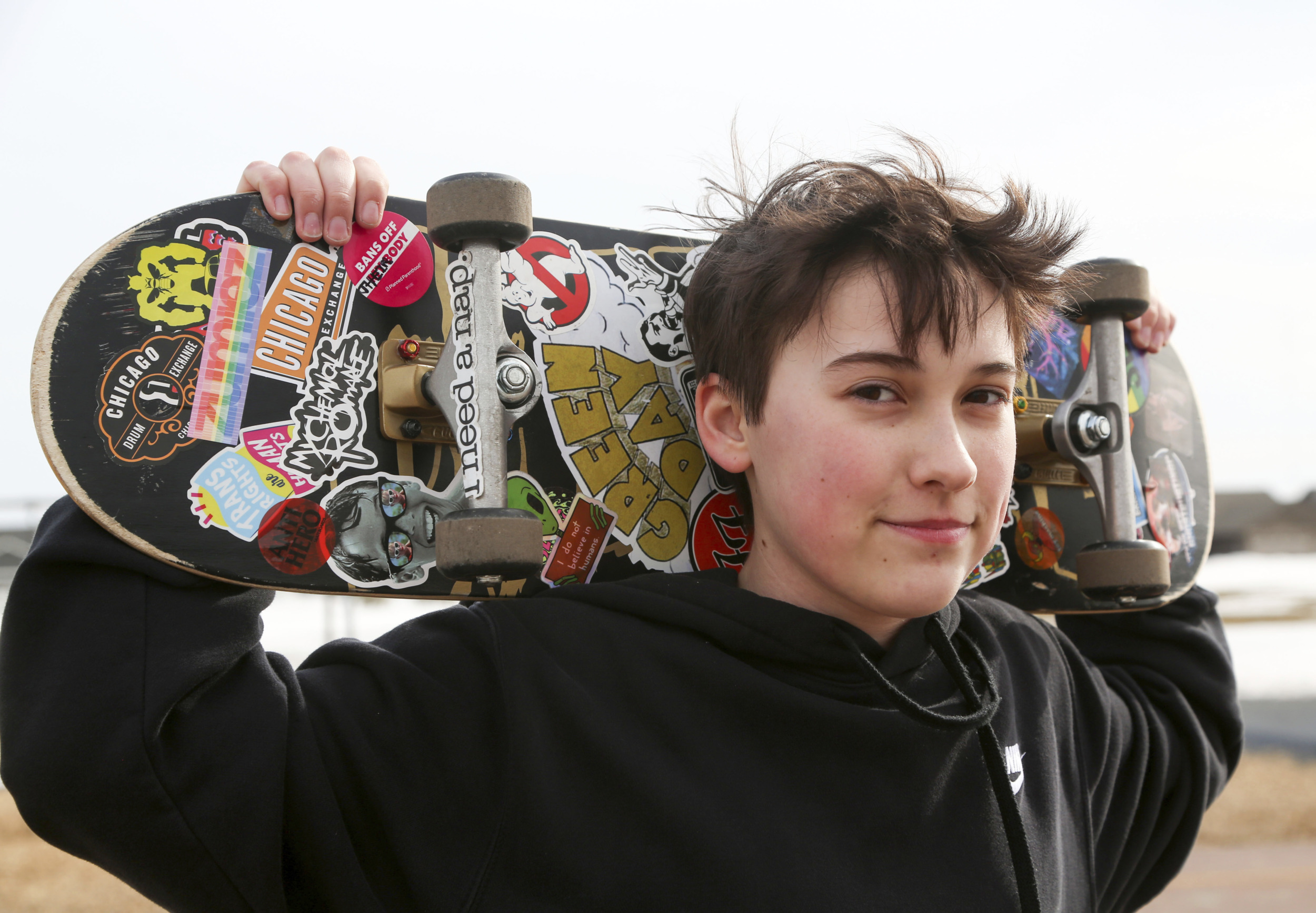Transgender treatment for kids: Young peron with shprt dark hair on black sweatshirt stands holding colorful skateboard behine their head with both hands.