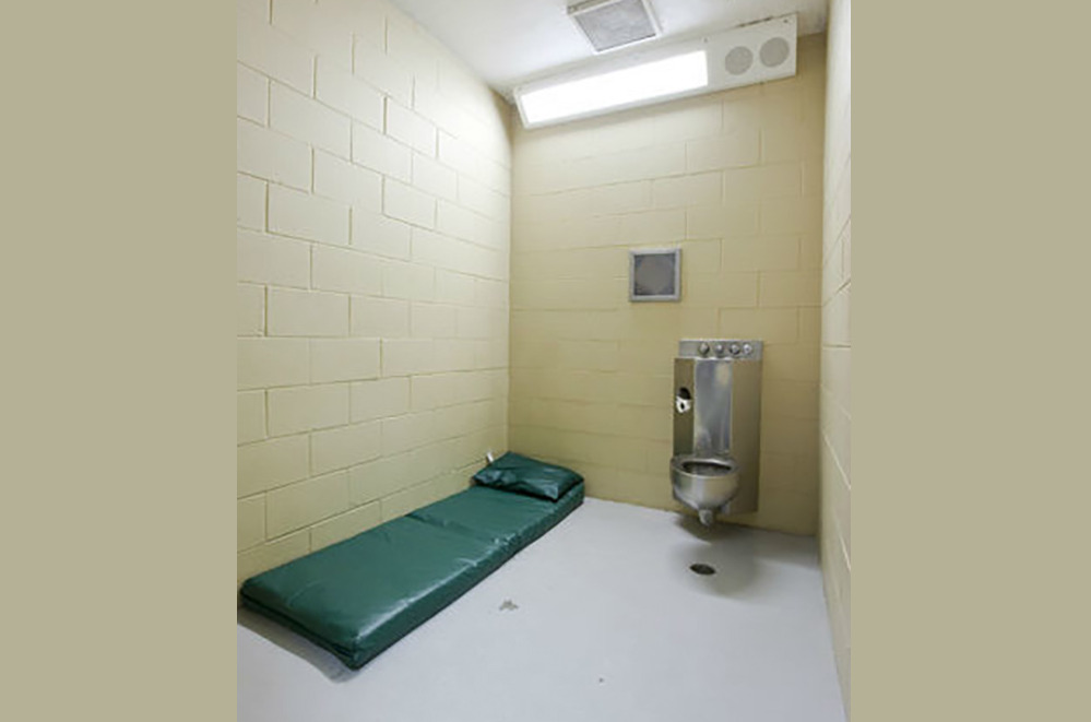 Juvenile solitary: Youth in dark, loose pants & shirt stands with back to camera in small white cell with built-in cement bed.