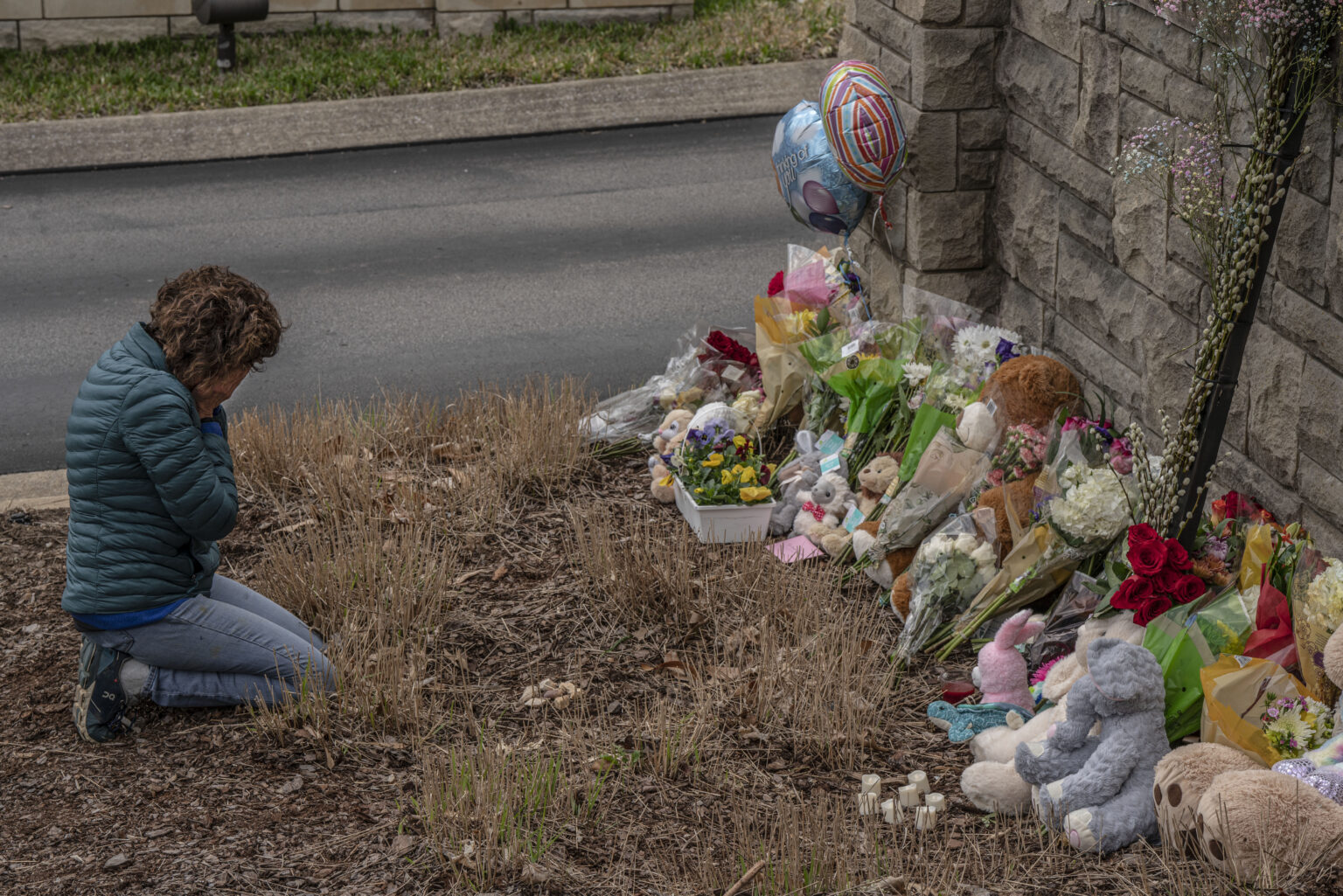 Tennessee School Drills: Person with short brown hair with head down and hands covering face kneels in dry grass in front of stone wall with flowers, suffed animals and balloons piled up against it