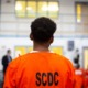 Youth In Adult Prisons: young black prisoner with back to camera in orange prison uniform looking at white wall of doors and interior windows