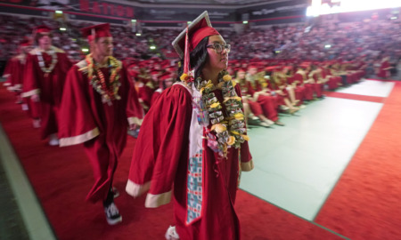 Teen Native American girl in red graduation cap and gown walks toward front of auditorium with seated fellow graduates in the background
