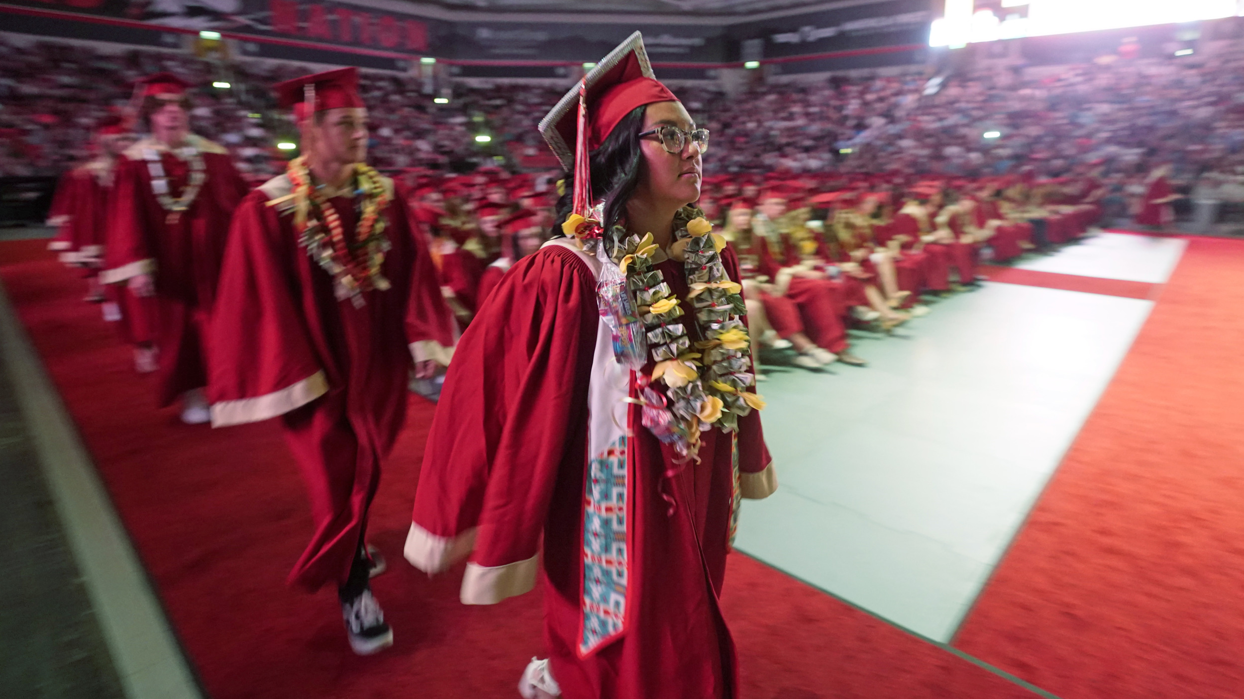 Teen Native American girl in red graduation cap and gown walks toward front of auditorium with seated fellow graduates in the background