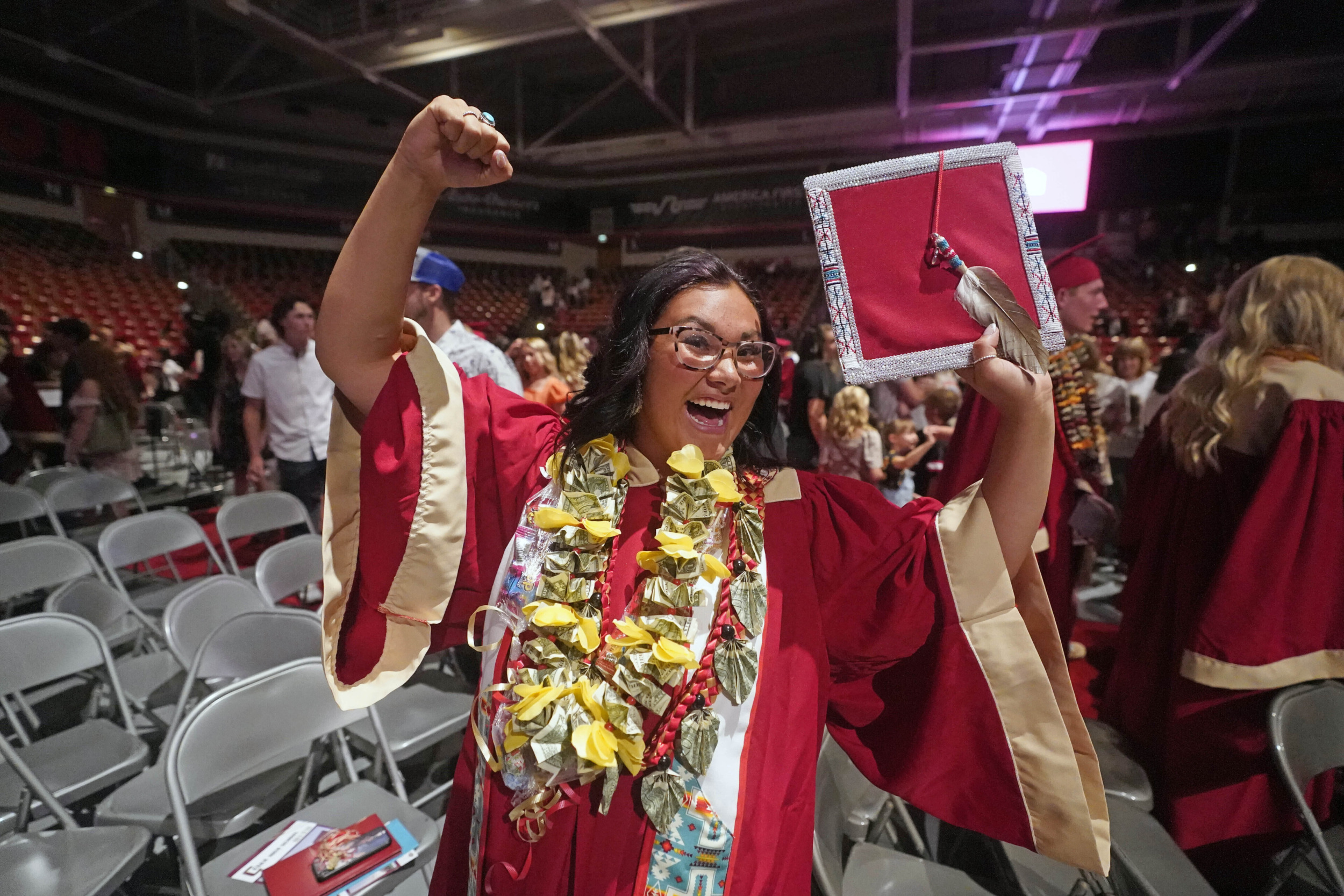 Indigenous Graduation Regalia: Young Native American teen in red cap and graduation robe raises hands with big smile on her face after receiving diploma.