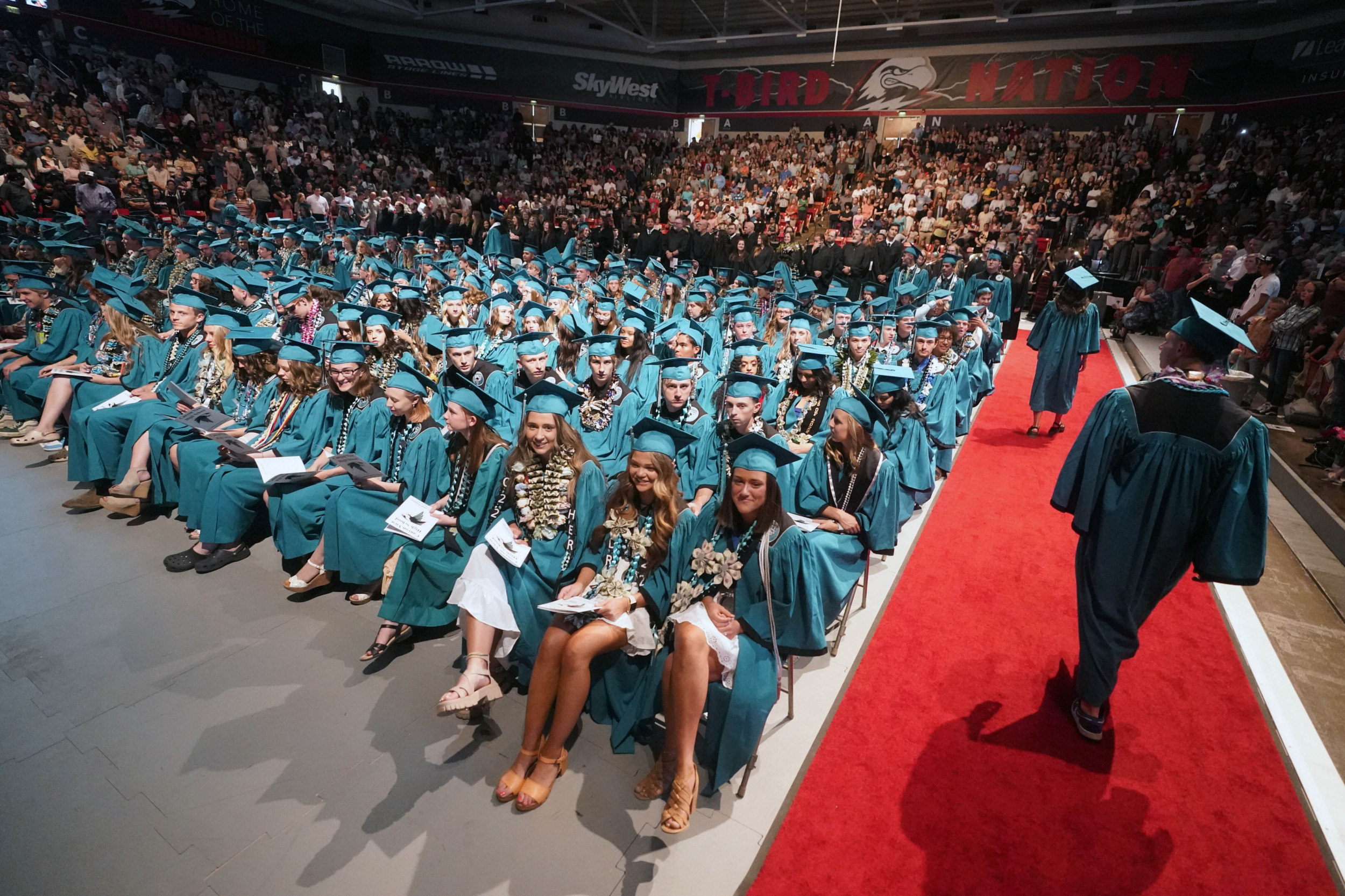 Indigenous Graduation Regalia: Large auditorium is filled with seated graduates wearing turquoise graduation caps and downs.