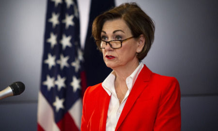 Child Labor Laws: Iowa Governor Kim Reynolds, a woman with short dark hair and dark-framed glasses, wearing red suit with white blouse, standing next to American flag while speaking into microphone.