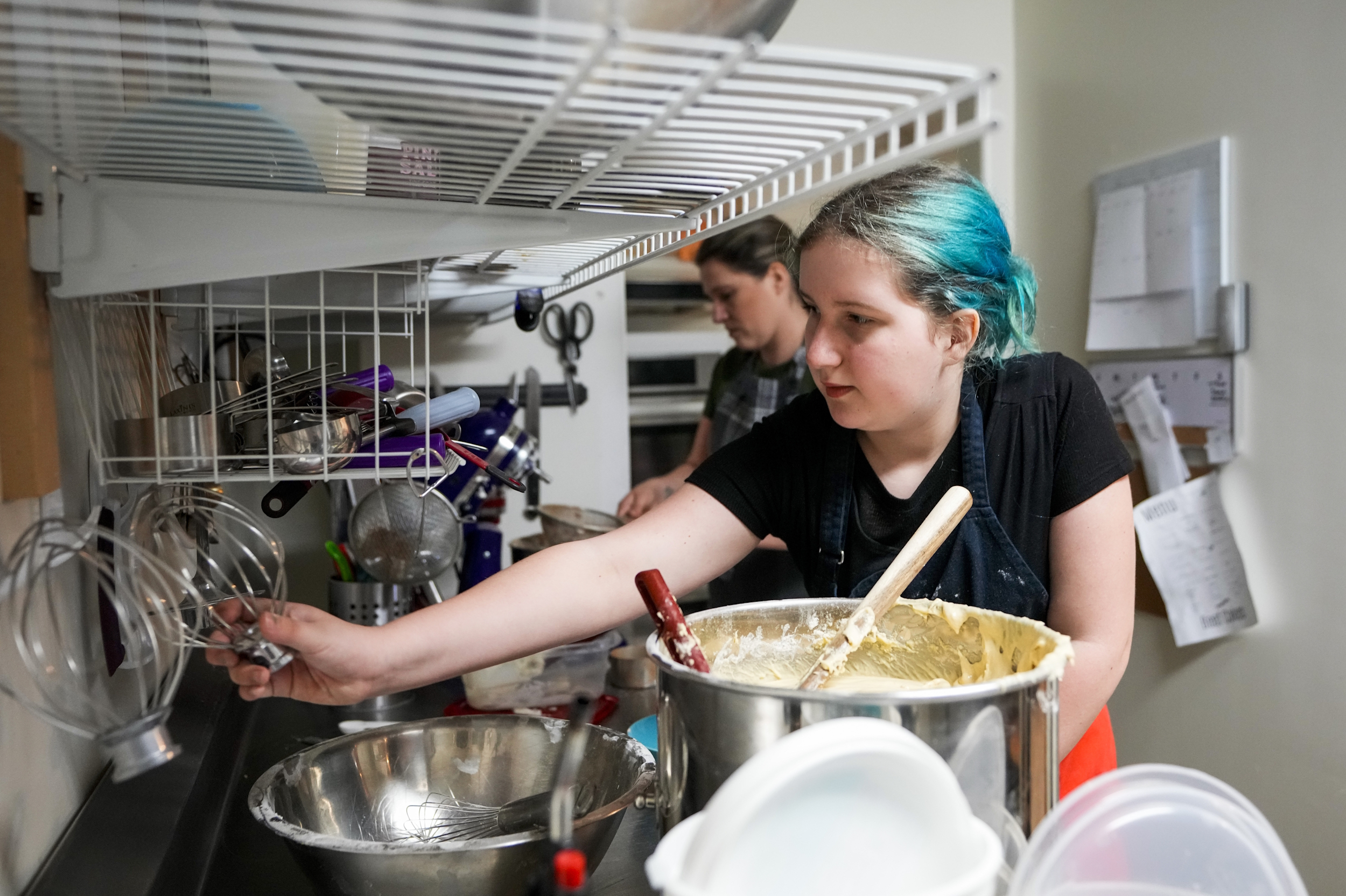 Disabled students' shorter school days: Teen girl with turquoise hair pulled back wearing black t-shirt reaches across several stainless steel mixing bowls for baking tools hanging on the wall in a commercial kitchen.
