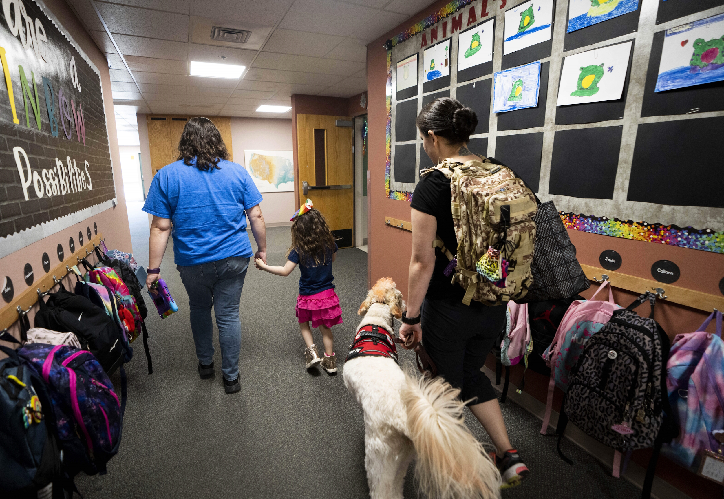 Disabled students' shorter school days: Woman in blue t-shirt and blue jeans holds hand of young girl wit dark hair in black top and pink skirt walking away from camera in school hallway followed by dark-haired woman with daypack leading large golden-coated dog on leash with red service dog vest..
