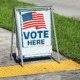 voting sign on sidewalk: "Vote Here" and American flag graphic on folding sidewalk sign