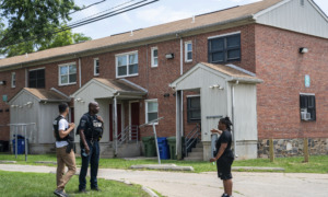 Mass shooting Baltimore: Black policeman in dark uniform stands with 2 black adults outside on grass in front of two-story, red brick apartment building with white porch shelters & trim.