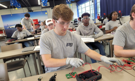 Flight Path 9 airbus training: High school teen in gray t-shirt with short blonde hair & glasses wearing protective gloves sirs at sesk working with electrical wiring equipment. Several students in background are doing the same.