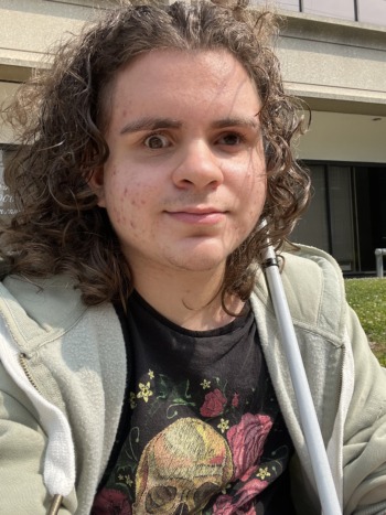 A selfie shot taken by EJ Valez with shoulder length curly hair, wearing a black t shirt with a skull stitched in colorful thread and a gray hoodie