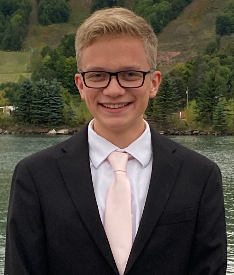 LGBTQ+ youth: Young blonde man with short hair and black-framed glasses in black suit, white shirt and pink tie with river and greenery in the background..