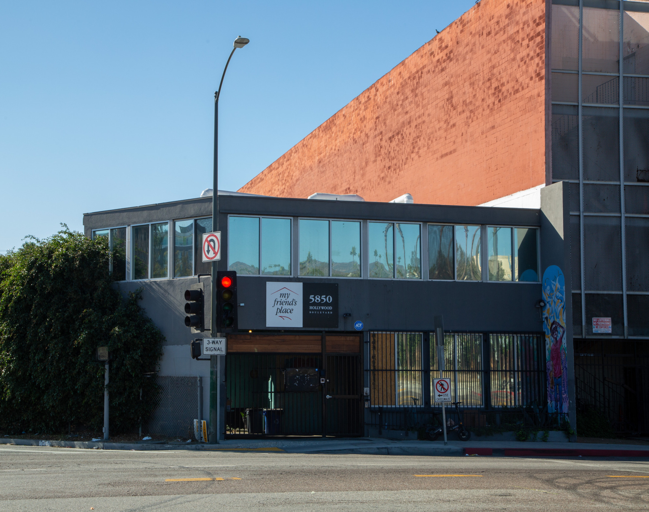 A view of My Friend's Place in Los Angeles, California. A two-story dark gray standard looking office block sits before a pale blue sky and a red brick building.