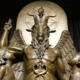 Satan Afterschool: Close ip of bronze stature of Baphimet, a traditional depiction of satan with horned goat head and wings in front of pentagram with right hand pointing to sky. A young boy and girl stand on either side of his knees looking up to him.