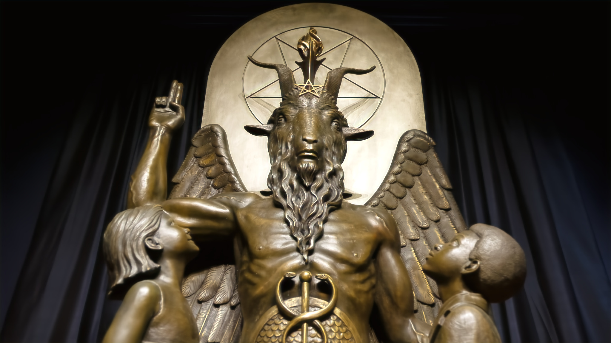Satan Afterschool: Close ip of bronze stature of Baphimet, a traditional depiction of satan with horned goat head and wings in front of pentagram with right hand pointing to sky. A young boy and girl stand on either side of his knees looking up to him.