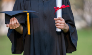 migrant student, college and university, higher education: Closeup of person in black graduation gown holding a black graduate cap with gold tassel and a rolled parchment scroll with red ribbon