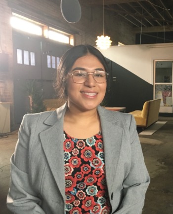 Juvenile justice reform: Young adult Latina woman with black hair pulled back and rimless glasses in light gray suit jacket with red print top stands in warehouse smiling into camera.