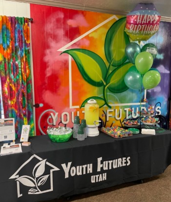 A rainbow bedecked promotional table at Youth Futures Utah, a shelter in Ogden, Utah. Their logo, a leaf in a house icon, is stamped on posters and other memorabilia on the table.