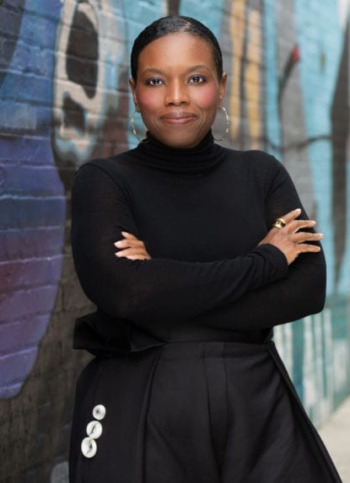 Young people lead us to a new vision of justice: Candice C. Jones - Black woman with black hair pulled back in black turtleneck and black pants stands with arms crossed in front of alleyway wall with brightly colored blue and black graffiti.
