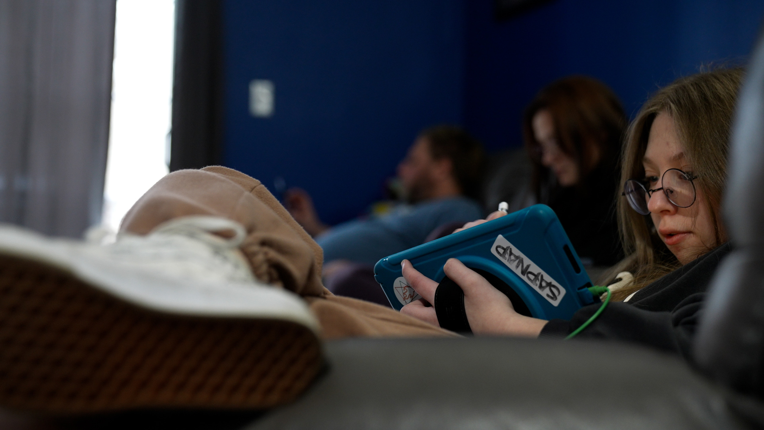 Long COVID — A girl with round glasses and long brown hair sits on the couch in her home and uses an iPad in a blue one-handed case.