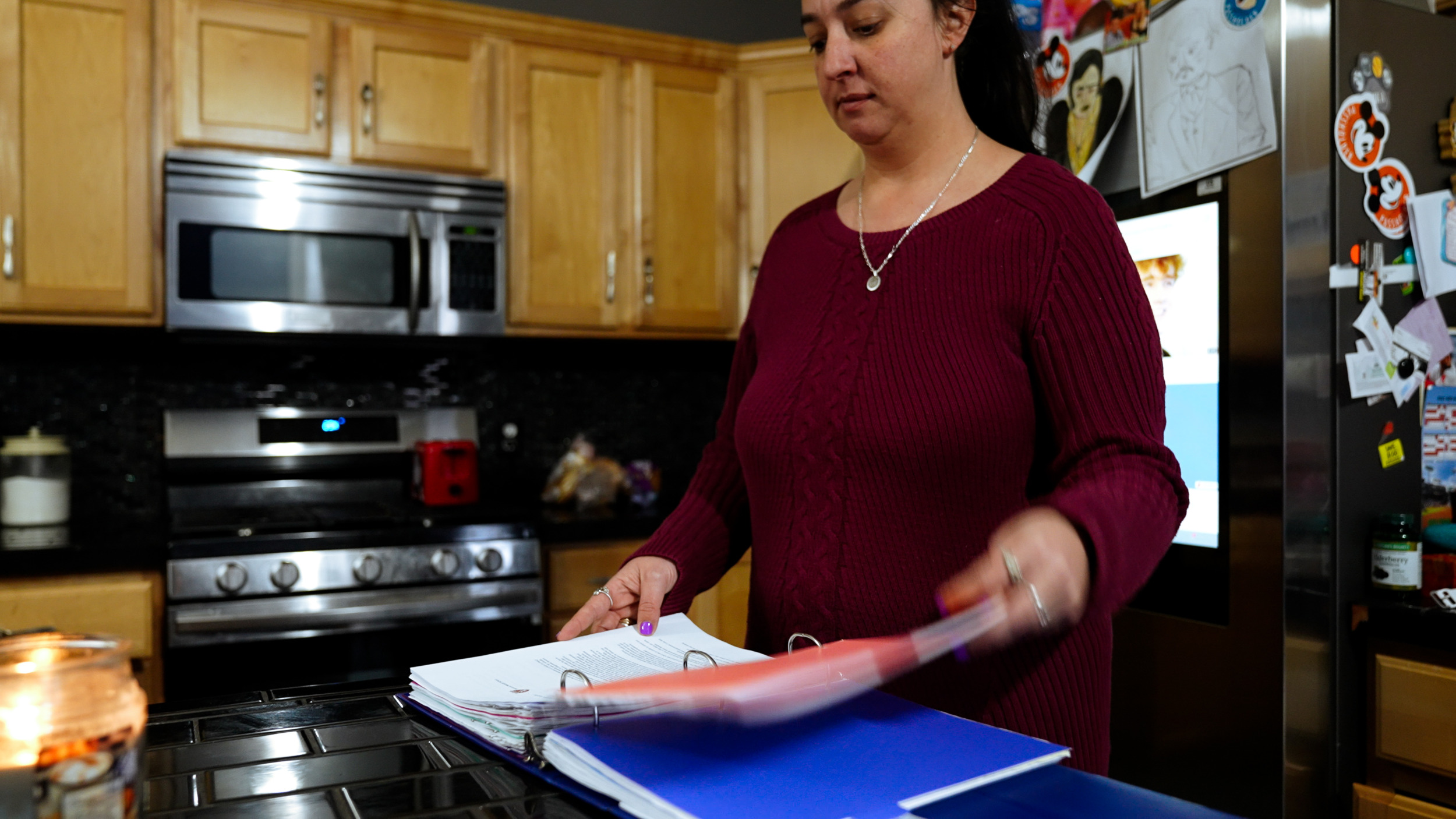 Long COVID — A woman with black hair wearing a silver chain with a round pendant on a maroon cable knit style sweater stands in front of kitchen island paging through a thick binder with many sections and papers.