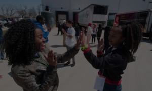 Youth-led project grants, community health, education equity, environmental justice, civics: two girls playing patty-cakes outside among larger group of youths