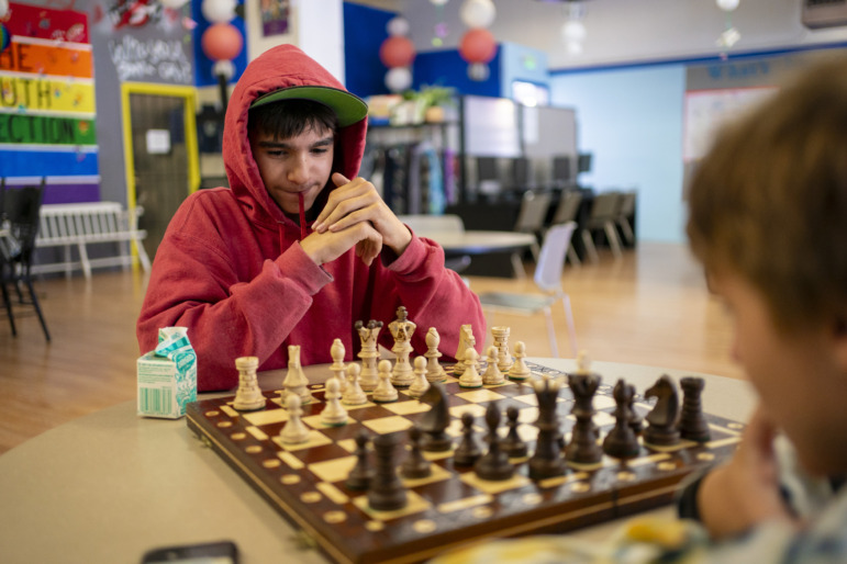 Homeless kids: A person with dark hair and a red hoodie sits with hands steepled behind a chess board
