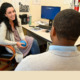 Job Psychologist: Woman with long. dark hair sits next to an office desk facing toward a Black person with short black hair
