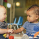 Home-based childcare business: Two young infants sit on floor side-by-side holding hands looking into each others eyes.
