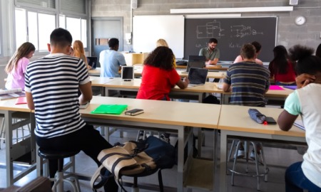 Classroom disruptions: Rear view of several students at desks bent over work with teacher facing classroom standing in front of blackboard.