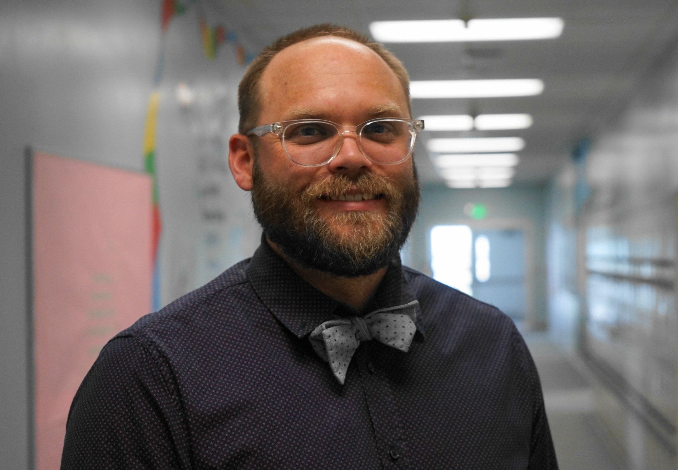 Chugwater, WY public school is now charter: bearded man with glasses and bowtie stands and smiles in school hallway