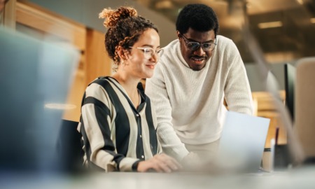Low-income community development grants: black man with glasses and white sweater helping young woman with curly hair and glasses on computer