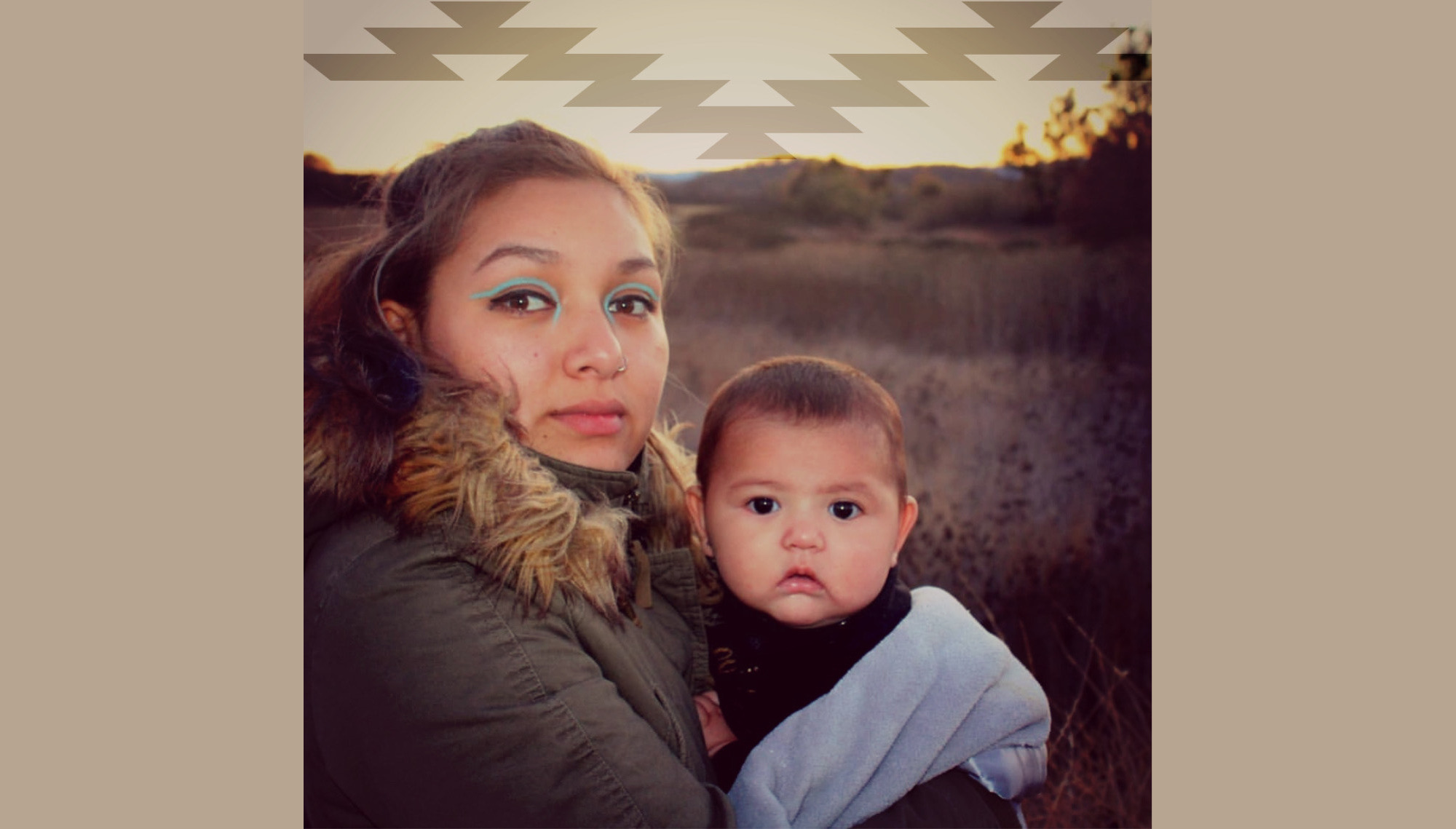 Wiyot Foster Youth Housing: Woman with long brown hair in brown winter coat with large fur collar stands outside in a filed of brown grass looking into camera holding infant draped in light blue blanket in her arms