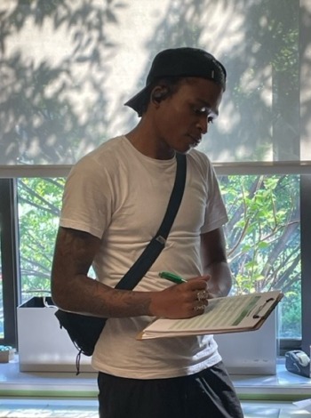 First-time juvenile offenders: Young adult Black man in white t--shirt with dark crossbody bag, gold rim glasses and dark cap stands in front of large window with view of greenery writing on papers on a clipboard