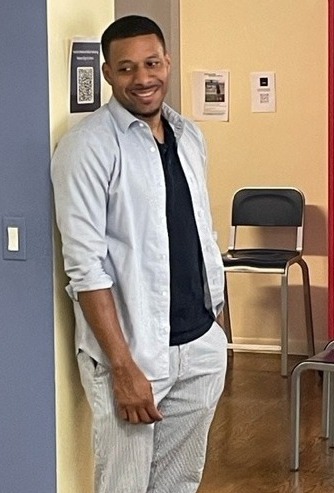 First-time juvenile offenders: Black man with short black hair and beard wearing white pant and shirt stands in room with chairs leaning against yellow wall