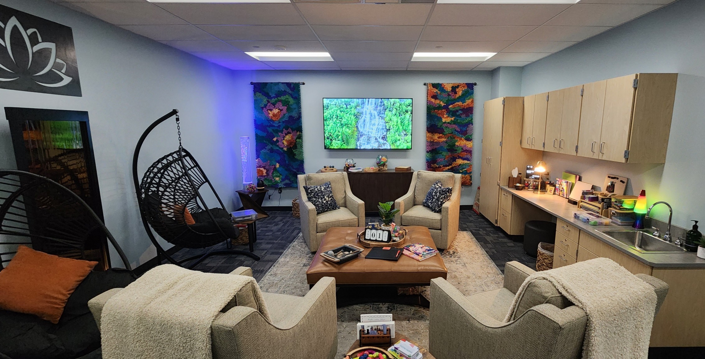 Stydent Wellness Center: A room furnshed like a cozy licing room with four overstuffed chairs around a large coffee table, a large screen TV, and colorful, large tactile art on the wall.