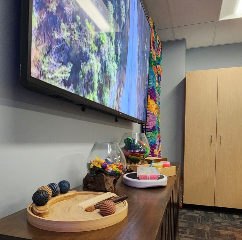 Student Wellness Center: Long, wood side table holds many types of fidget widgets, with a large video screen above the table, and a bright, multicolor tactile, interactive art tapestry hangd on the wall.