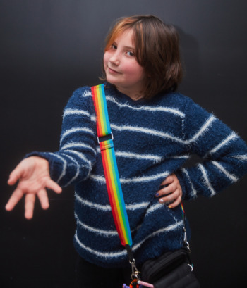Youth podcaster: Young preteen with bobbed brown hair wearing navy and white stripe sweater with rainbow strap cross-body bag stands with hand on hop and other hand splayed palm reaching toward camera