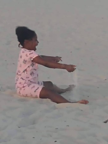 Guaranteed income: Young black girl in pink print top and shorts sits on white sand beach plating in the sand.