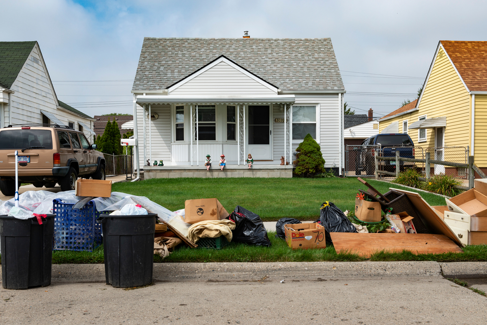 Youth Eviction Homelessness: Long pile of trash and two full trash cans cover the curbside grass in front of a small, white clapboard house with three dolls sitting on edge of porch.