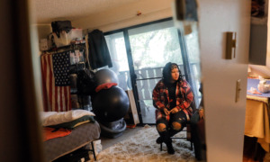Washington homelessness and foster care: Young woman with long, dark, curly hair in red and black plaid jacket, black top and ripped jeans, sits in office chair in room with large sliding glass door.