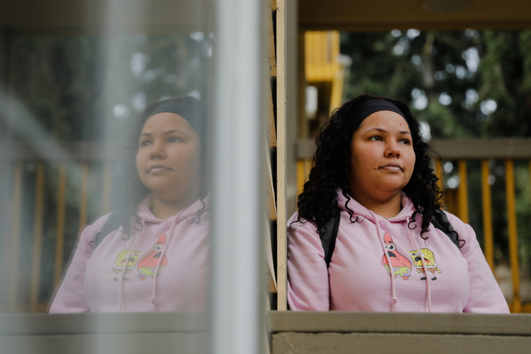 Washington homelessness and foster care: Young woman with long, dark, curly hair in pink sweatshirt stares pensively into the distance standing outside next to a building.