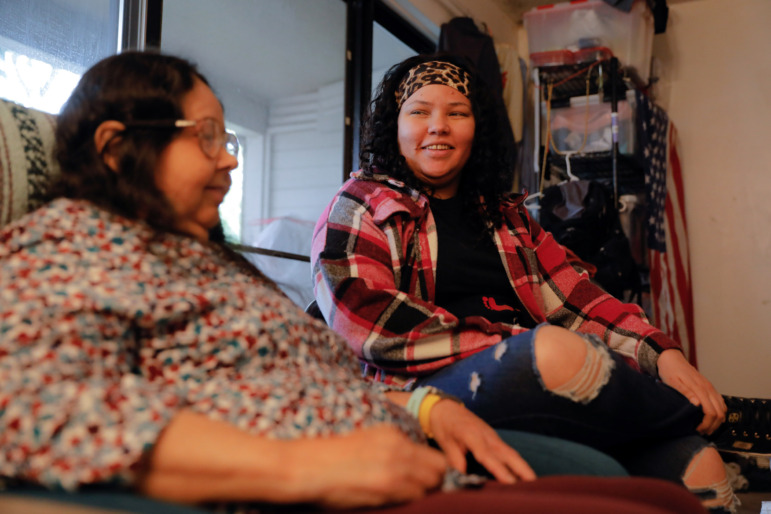 Washington homelessness and foster care: Young woman with long, dark, curly hair in red and black plaid jacket, black top and ripped jeans, sits next to older dark-haired women with glasses and multi-colored print top in front of large window.