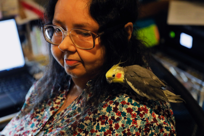 Washington homelessness and foster care: Close-up of middle-aged, dark-haired women with glasses and multi-colored print top sits with pensive look while a small gray bird with yellow head sits on her shoulder.